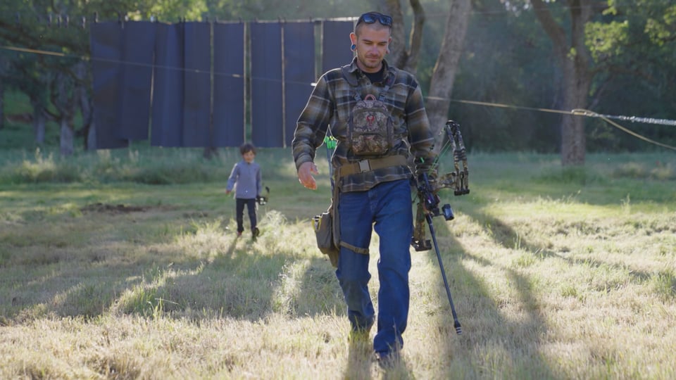 Man walking through a field carrying bowhunting gear, with a child following behind