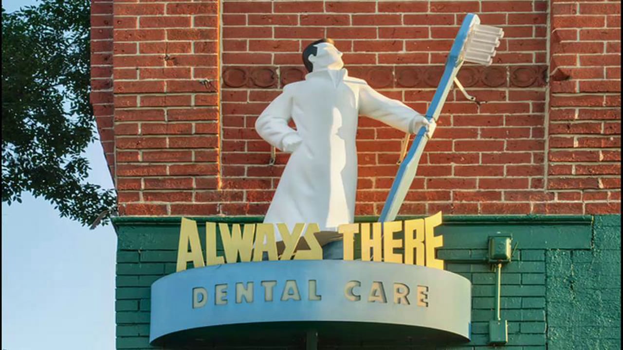 The Always There Dental Care storefront sign, a white statue of a dentist holding a giant blue toothbrush above the practice name