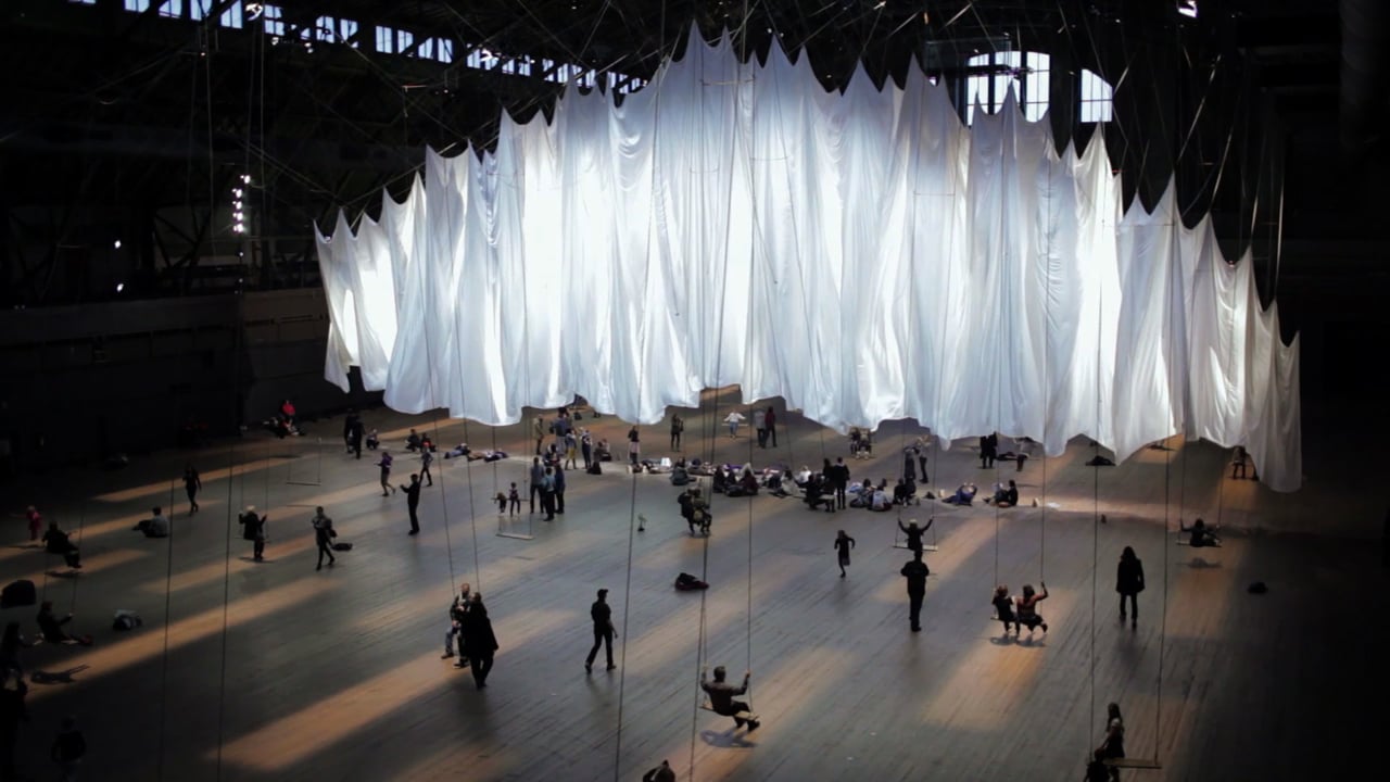 Ripples of white sheets arc from the ceiling. On the ground, clear strings hold up various figures.